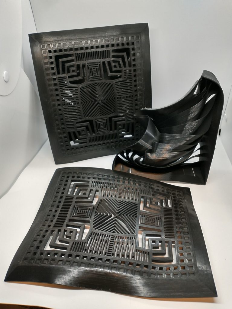 Three components of two grates, all with various levels of heat distortion. In the foreground is the front face of one grate with significant warping. in the mid right there is the box for the grate, severely collapsed, the vent vanes folded like ribbons. In the back is an intact assemblybut many parts of the design are warped out of shape.