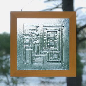 A square wooden frame contains a panel of textured glass. Several rectangular pieces of glass are fused to the panel to form a geometric pattern.