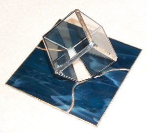 A cube made from transparent glass appears to be embedded in a square of blue glass with white streaks.